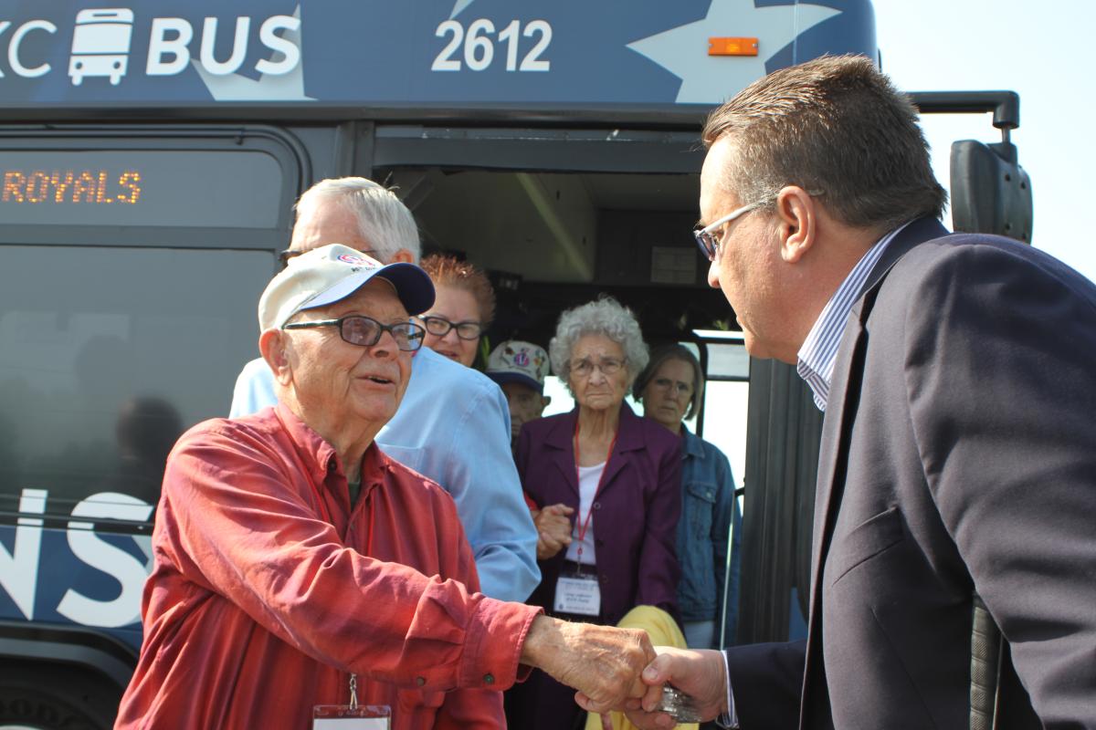 Group of older people exiting a bus. Man in glasses at front of line shaking hands with man in suit wearing glasses